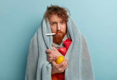 Redhead young man with bristle fights against flu and fever, holds lemon to enrich vitamins, has thermometer in mouth, trembles under blanket, isolated over blue background. Health problems.