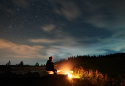 Tourist man sitting near cozy bonfire and relaxing on fresh air. Concept of good weather and process enjoying incredible starry sky at night.