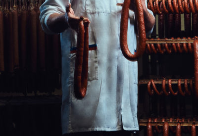 Handsome butcher wearing uniform holding sausages in the storage room.