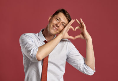 Studio portrait of a good-looking young guy in a classic white shirt and red tie, showing a heart folded of his fingers and smiling while posing over a pink background. Stylish haircut. Sincere emotions concept. Copy space.