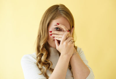 Isolated studio shot of glamourous stylish young European female with red manicured nails covering her face with hands and peeping through her fingers at camera, feeling shy, ashamed or scared